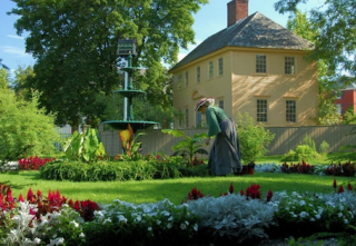 Large flower garden with a fountain and a woman dressed in Victorian-era clothing, with large house in background